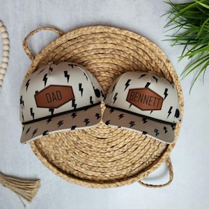 May include: Two beige trucker hats with black lightning bolt patterns. One hat has a brown leather patch that says "DAD" and the other has a brown leather patch that says "BENNETT".