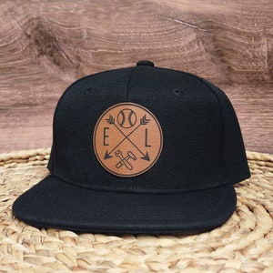 May include: Black baseball cap with a brown leather patch featuring a baseball, crossed arrows, and the letters "E" and "L".