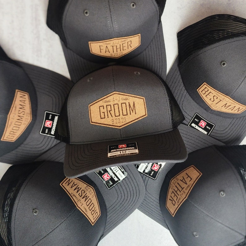 May include: A collection of gray trucker hats with black mesh backs. Each hat features a tan leatherette patch with text such as "GROOM", "FATHER", and "BEST MAN", along with a date. The hats are from the brand Richardson.