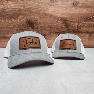 May include: Two gray and white trucker hats with brown leather patches. The larger hat has the word "Dad" on the patch. The smaller hat has the name "Bennett" on the patch.