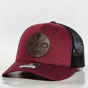 May include: A maroon and black trucker hat with a brown leather patch featuring a cross, a football, the number 11, and the word "SMITH".