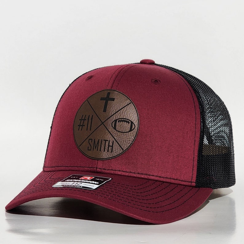May include: A maroon and black trucker hat with a brown leather patch featuring a cross, a football, the number 11, and the word "SMITH".