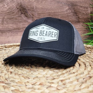 May include: A black trucker hat with a gray mesh back and a curved brim. The hat features a white patch with the words "RING BEARER" and the date "02.18.24" in black text. The hat is sitting on a woven surface.