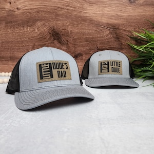 May include: Two gray and black trucker hats with brown leather patches. The larger hat has the text "Little Dude's Dad" on the patch. The smaller hat has the text "Dad's Little Dude" on the patch.