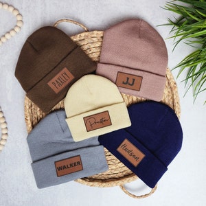 May include: Five different colored knit beanies with personalized leather patches. The beanies are brown, pink, cream, gray, and navy blue. The patches have the names "Paxley", "JJ", "Preston", "Walker", and "Hudson" written on them.