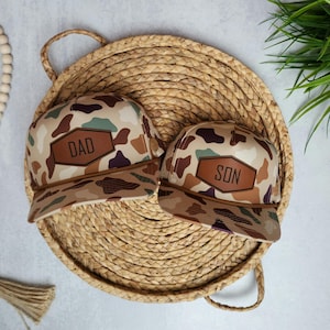 May include: Two camouflage trucker hats with brown leather patches. The larger hat has the word "DAD" on the patch and the smaller hat has the word "SON" on the patch.