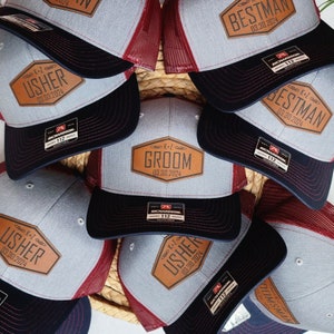May include: A collection of gray and black trucker hats with leather patches. The patches feature the words "Groom," "Bestman," and "Usher," along with a date. The hats have a mesh back and a Richardson 112 tag.
