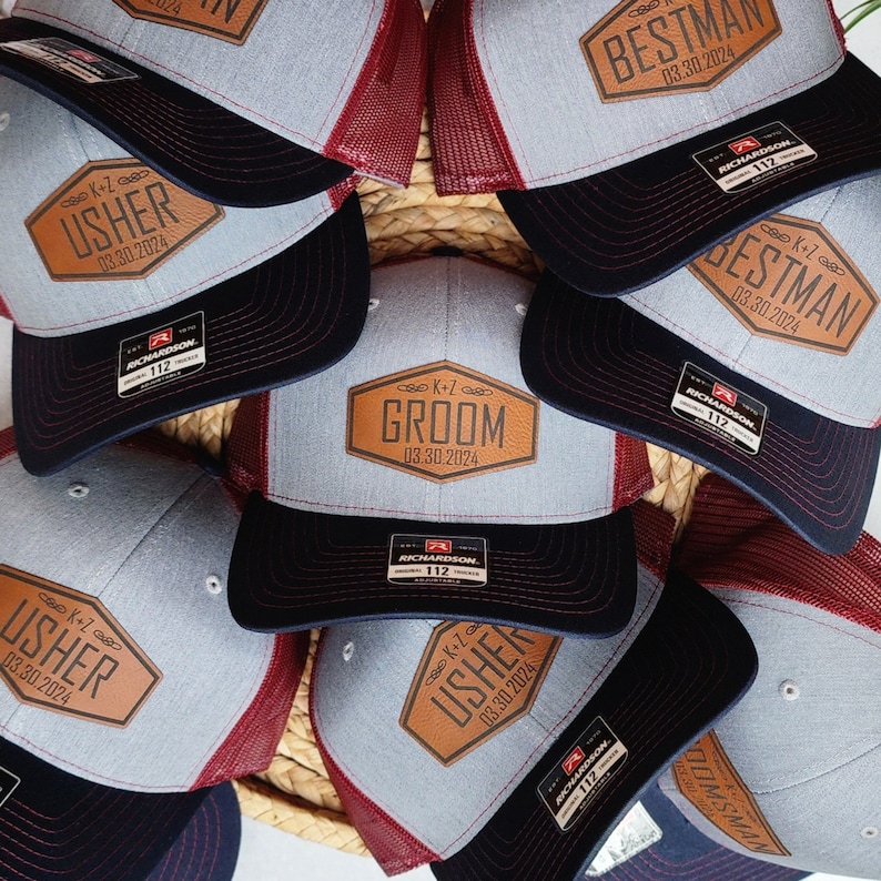 May include: A collection of gray and black trucker hats with leather patches. The patches feature the words "Groom," "Bestman," and "Usher," along with a date. The hats have a mesh back and a Richardson 112 tag.
