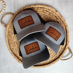 May include: Three gray and black trucker hats with brown leather patches. The patches have the names "Dada", "Kenny", and "Kaiden" written in cursive.