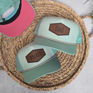 May include: Three trucker hats with a light green and brown color scheme. Two hats have brown leather patches with the names "DAD" and "WALKER". The third hat has a pink underside and a light green top.