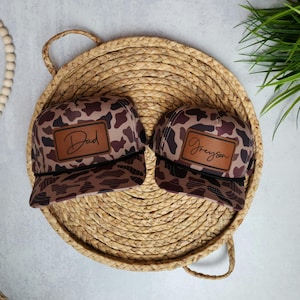 May include: Two camouflage trucker hats with brown leather patches. The patch on the left hat says "Dad" and the patch on the right hat says "Greyson".