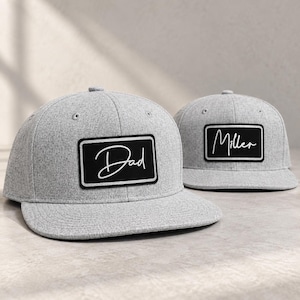 May include: Two heather gray baseball caps with flat brims. Each cap features a rectangular black patch on the front. One patch displays the word Dad in white cursive script, and the other patch displays the word Miller in white cursive script.