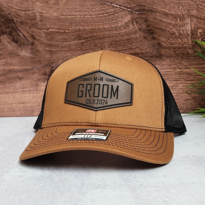 May include: A brown trucker hat with a black mesh back and a curved brim. The front features a leather patch with the word "GROOM" and the date "05.11.2024". The hat has an adjustable snap closure.