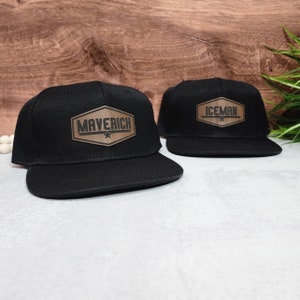 May include: Two black baseball caps with brown leather patches. The patch on the left cap says "Maverick" and the patch on the right cap says "Iceman".
