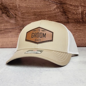 May include: Tan and white trucker hat with a brown leather patch. The patch features the word "GROOM" in black lettering, along with the date "8.4.23" and the initials "CG". The hat has a curved brim and a mesh back.