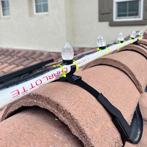 May include: Close-up of a white Christmas light string with clear bulbs attached to a roof using black straps. The string is labeled "CHARLOTTE" and "LED LIGHT 6641 100".