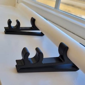 May include: Two black plastic window blind brackets mounted on a white window sill. The brackets are designed to hold a white window blind rod.
