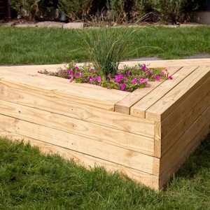 May include: A triangular wooden planter box with pink flowers and green plants. The planter is made of light-coloured wood and sits on green grass. The design is modern and suitable for outdoor use.