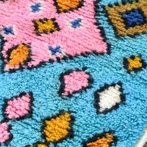 K&ouml;nnte beinhalten: Nahaufnahme eines bunten, strukturierten Teppichs mit geometrischem Muster. Der Teppich zeigt Farbt&ouml;ne von Blau, Rosa, Gelb und Braun.