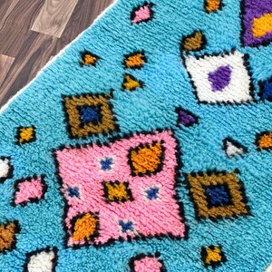 K&ouml;nnte beinhalten: Ein blauer Teppich mit einem geometrischen Muster in Rosa, Orange, Gelb, Wei&szlig; und Lila. Der Teppich hat eine flauschige Textur.