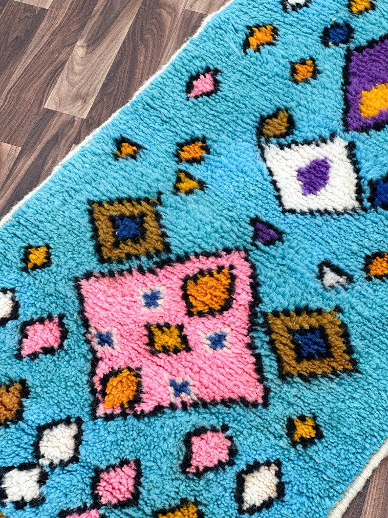 K&ouml;nnte beinhalten: Ein blauer Teppich mit einem geometrischen Muster in Rosa, Orange, Gelb, Wei&szlig; und Lila. Der Teppich hat eine flauschige Textur.