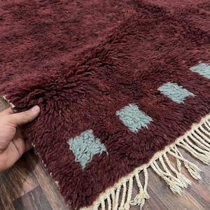 May include: A burgundy-colored, textured rug with a shaggy pile. The rug features a pattern of light blue square shapes and a fringe border. The rug is made of a soft material and is suitable for home decor.