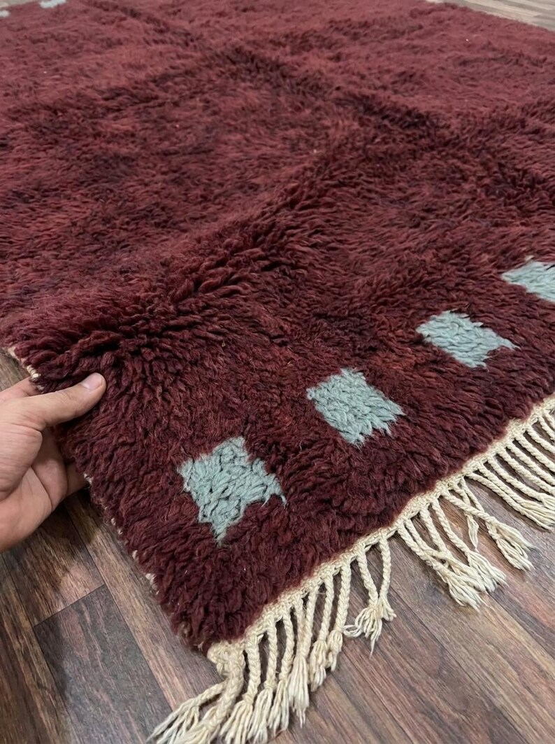 May include: A burgundy-colored, textured rug with a shaggy pile. The rug features a pattern of light blue square shapes and a fringe border. The rug is made of a soft material and is suitable for home decor.