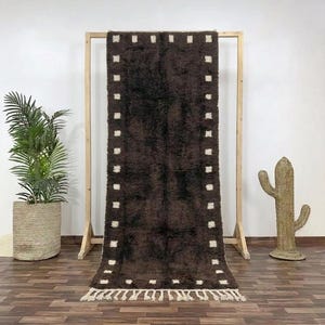 May include: A dark brown rectangular rug with a shaggy texture and a border of white square accents. The rug is displayed on a wooden rack, showcasing its design and texture. The rug has a fringe at the bottom.