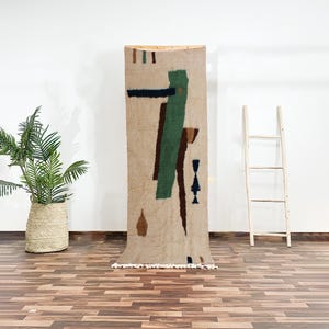 Op de afbeelding: Een beige wollen tapijt met abstracte geometrische vormen in groen, bruin en zwart. Het tapijt hangt aan een muur en heeft een franje rand.