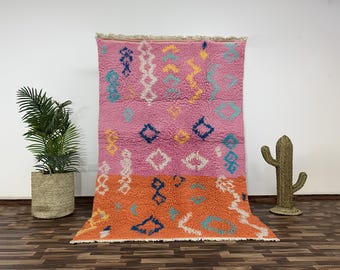 May include: A large, rectangular rug with a pink and orange color scheme. The rug features geometric patterns in white, blue, and yellow. The rug is made of a thick, textured material, and has a fringed edge. The rug is displayed against a white wall.