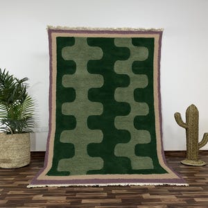 Könnte beinhalten: Ein rechteckiger grüner Teppich mit einem sich wiederholenden hellgrünen Muster. Der Teppich hat einen beigen Rand mit lila Einfassung. Eine Topfpflanze und eine Kaktusdekoration befinden sich im Hintergrund.