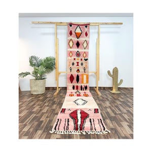 Peut inclure: Un long tapis de couloir rose et crème avec des motifs géométriques rouges, noirs et orange. Le tapis est exposé sur un support en bois, mettant en valeur son design et sa texture complexes. Le design du tapis comprend des losanges et des rectangles.