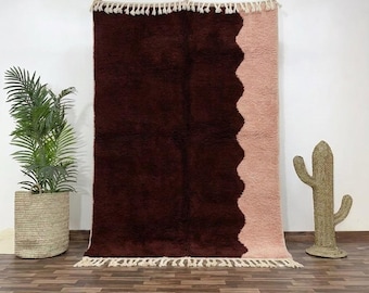 May include: A large, rectangular rug with a dark burgundy field and a light pink border with a wavy edge. The rug has a shaggy texture and is adorned with cream-colored fringe along the top and bottom edges. A decorative cactus and potted plant are in the background.