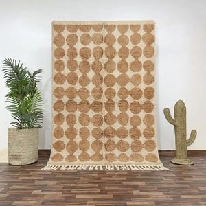 May include: A large, rectangular rug with a repeating pattern of brown circles on a cream background. The rug has a fringed edge and is displayed against a white wall. A potted plant and a cactus decoration are on either side.