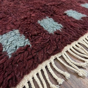 May include: Close-up of a burgundy rug with a geometric pattern of light blue squares. The rug has a thick, textured pile and a cream-colored fringe along the edge. The rug is a decorative home accent.