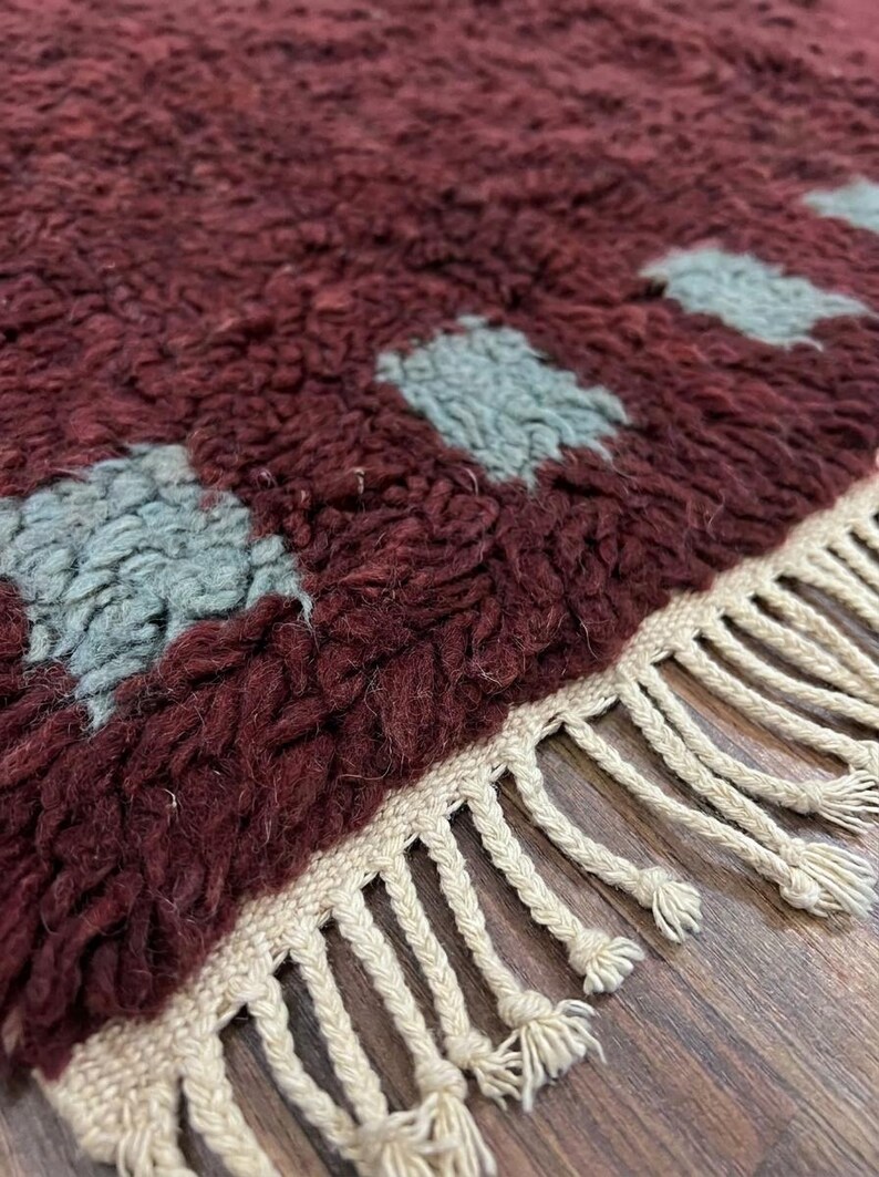 May include: Close-up of a burgundy rug with a geometric pattern of light blue squares. The rug has a thick, textured pile and a cream-colored fringe along the edge. The rug is a decorative home accent.