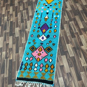 K&ouml;nnte beinhalten: Ein langer, rechteckiger Teppich mit blauem Hintergrund und einem farbenfrohen geometrischen Muster. Das Muster enth&auml;lt rosa, gelbe, wei&szlig;e und violette Rauten und Dreiecke.