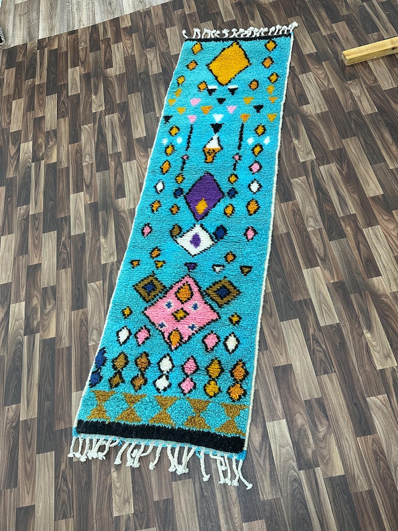K&ouml;nnte beinhalten: Ein langer, rechteckiger Teppich mit blauem Hintergrund und einem farbenfrohen geometrischen Muster. Das Muster enth&auml;lt rosa, gelbe, wei&szlig;e und violette Rauten und Dreiecke.