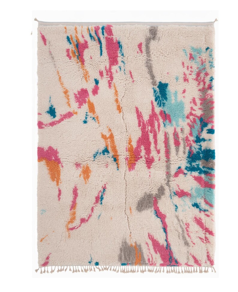 May include: A rectangular rug with a cream-colored background and a colorful abstract design. The design features splashes of pink, orange, teal, and gray. The rug has a fringed edge and is made of a soft, textured material.
