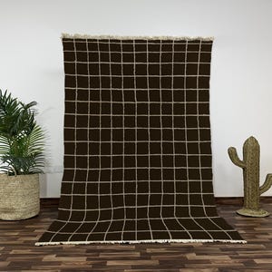 Peut inclure: Un tapis marron foncé avec un motif quadrillé de lignes beige clair. Le tapis a un bord effiloché et est présenté contre un mur blanc. Une plante en pot et une décoration de cactus sont de chaque côté du tapis.
