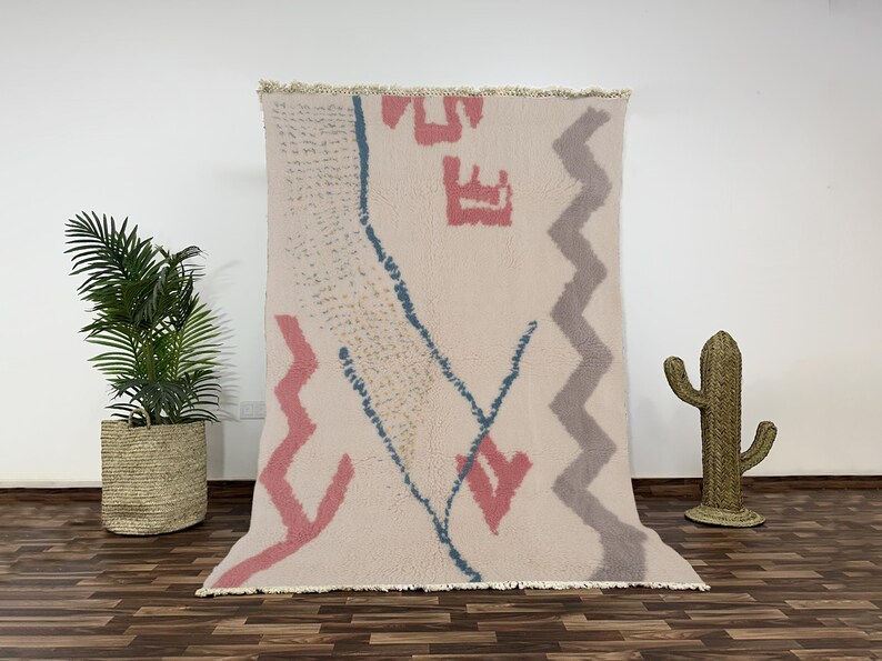 "A hand-knotted Moroccan wool rug with a plush cream pile, featuring modern abstract geometric shapes including pink and grey zig-zags and a speckled blue line, laid flat on a white background."