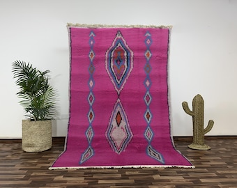 May include: A vibrant pink Moroccan rug with a central diamond pattern in blue, orange, and white. The rug features a repeating diamond design along the sides. The rug is displayed on a wooden floor.