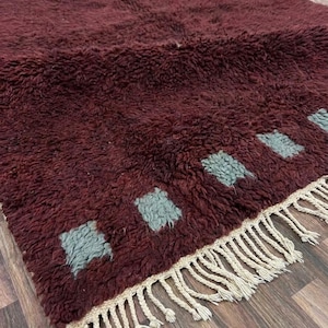 May include: A burgundy, shaggy rug with a geometric pattern of gray squares. The rug has a fringed edge in a natural beige color. The texture appears soft and plush, suitable for a living room or bedroom.