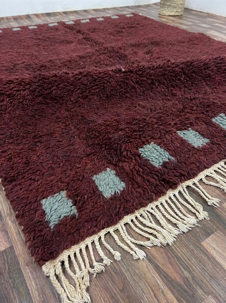 May include: A burgundy, shaggy rug with a geometric pattern of gray squares. The rug has a fringed edge in a natural beige color. The texture appears soft and plush, suitable for a living room or bedroom.