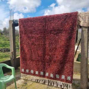 May include: A large, rectangular, burgundy-colored rug with a shaggy texture is hanging outdoors. The rug has a white border with small, light blue squares and white fringe. A green plastic chair is in the foreground.