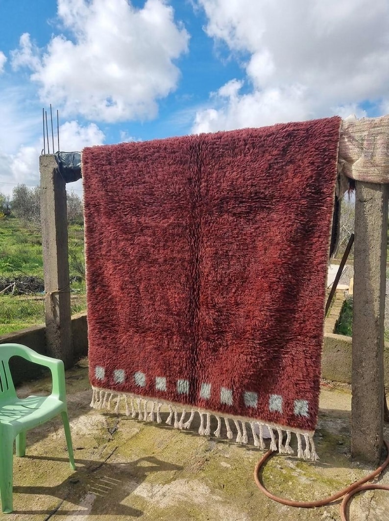 May include: A large, rectangular, burgundy-colored rug with a shaggy texture is hanging outdoors. The rug has a white border with small, light blue squares and white fringe. A green plastic chair is in the foreground.