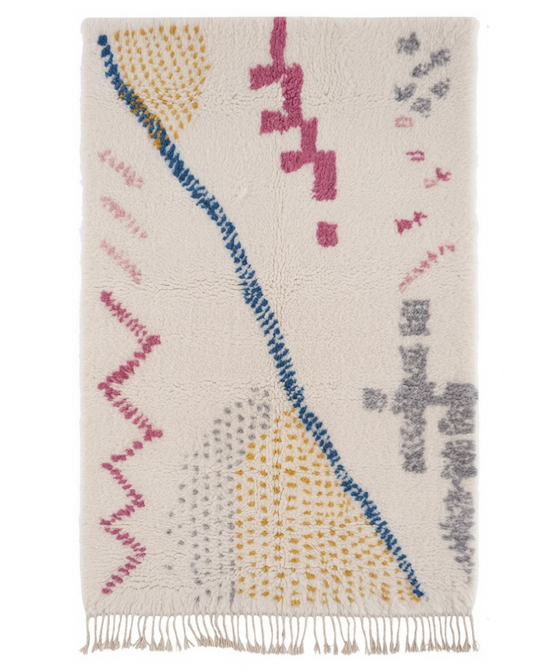K&ouml;nnte beinhalten: Cremefarbener rechteckiger Teppich mit geometrischem Design. Er hat eine blaue Diagonallinie, rosa Zickzackmuster und gelbe und graue gepunktete Muster. Der Teppich hat einen Fransenrand, der Textur und visuelles Interesse verleiht.