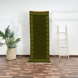 Op de afbeelding: Een rechthoekig olijfgroen tapijt met een donkerdere groene rand en een franje aan de onderkant. Het tapijt hangt aan een houten frame tegen een witte muur. Een potplant en een houten ladder staan ook in de afbeelding.