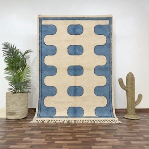 May include: A rectangular rug with a cream-colored background and a repeating pattern of blue, rounded rectangles. The rug has a blue border and fringe along the bottom edge. A potted plant and a cactus decoration are in the background.