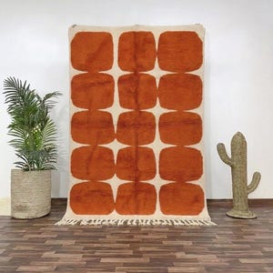 May include: A large, rectangular rug with a repeating pattern of rust-colored, rounded squares on an off-white background. The rug has a fringed edge and is displayed against a white wall. A potted plant and a cactus-shaped decor are on the floor.
